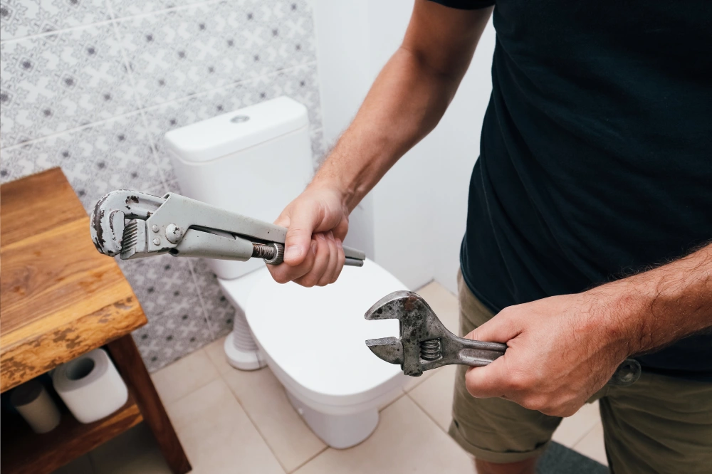 A man, a plumber, is holding two used adjustable wrenches in his hands while standing in a bathroom with a white toilet and patterned tiles in the background. Plumbing repair service.