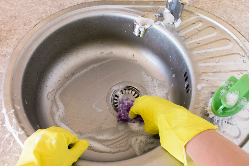 Woman in yellow rubber gloves washes and cleans a sink with a sponge and brush